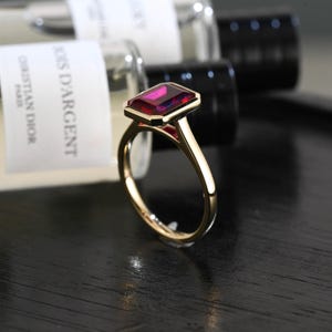 Op de afbeelding: Een gouden ring met een rechthoekige, smaragdgeslepen dieprode edelsteen gezet in een gepolijste metalen band. De ring staat rechtop en toont de levendige rode steen en het gladde, reflecterende oppervlak van de gouden band in een helder beeld.