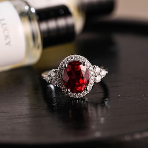 May include: A silver ring featuring a large oval ruby set within a halo of smaller diamonds. The band is delicate and adorned with additional diamonds on either side of the ruby.