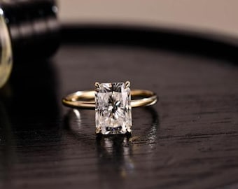 Puede incluir: Un anillo de oro con un diamante de talla rectangular. El diamante está engastado con cuatro garras, y la banda es sencilla. El anillo se presenta sobre una superficie oscura y texturizada, resaltando el brillo del diamante.