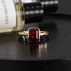 Puede incluir: Un anillo de oro con una piedra preciosa rectangular de color rojo intenso, tallada en forma de esmeralda y montada en un engaste liso. La banda es de oro pulido y la piedra muestra un tono carmesí saturado con facetas brillantes.