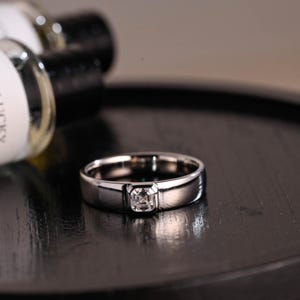 May include: A silver band ring with a square-shaped clear gemstone set in the centre.