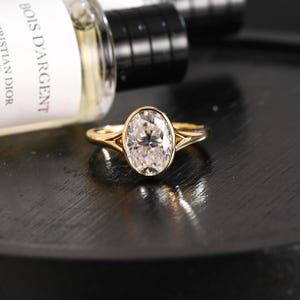Puede incluir: Un anillo de oro con una gran piedra preciosa transparente de talla ovalada y facetas, montada en un engaste de bisel. La banda tiene un diseño de vástago dividido que se estrecha hacia la piedra central. El anillo está frente a una botella Bois D'Argent.