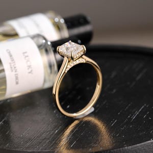 Peut inclure: Une bague de fiançailles en or avec une pierre centrale en diamant de taille carrée. La bague présente un anneau délicat et de petites pierres d'accent autour du diamant principal. La bague est présentée sur une surface sombre, avec des flacons de parfum flous en arrière-plan.