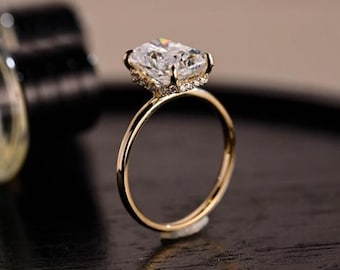Könnte beinhalten: Ein goldener Ring mit einem großen, klaren, rechteckig geschliffenen Edelstein als Mittelpunkt. Der Edelstein ist von kleineren, funkelnden Steinen umgeben. Der Ring ist aus poliertem Gold. Der Ring ist vor einem dunklen Hintergrund platziert.