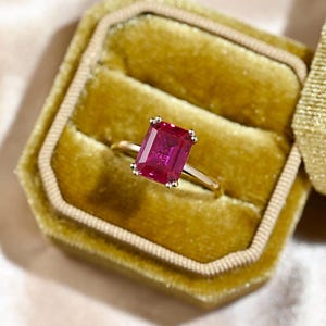 May include: A gold ring featuring a large, rectangular, bright pink gemstone. The ring is presented in a velvet box with a gold clasp. The gemstone's vibrant colour and cut are the focal point. The ring is a piece of jewellery.