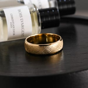 Può includere: Un anello color oro caratterizzato da una finitura testurizzata a linee incrociate sulla fascia esterna. L'anello presenta una superficie interna liscia e lucida e un design con bordi smussati. Il metallo riflette la luce, mettendo in risalto.