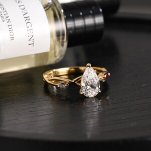 May include: A gold ring with a pear-shaped diamond as the centerpiece. The ring has a twisted band design with small accent stones, including a red ruby and two blue stones. The ring is set against a dark background.