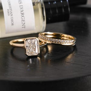 May include: A gold engagement ring featuring a rectangular radiant-cut centre stone in a bezel setting, paired with a matching gold wedding band encrusted with a row of small round white diamonds along the top surface of the metal band.