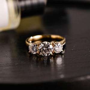 Può includere: Un anello con fascia in oro con tre pietre rotonde e trasparenti. L'anello è su una superficie nera.