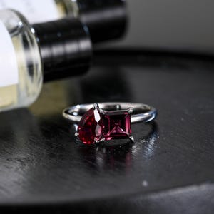 Può includere: Un anello d'argento con due pietre preziose rosse. L'anello ha una pietra preziosa a forma di pera a sinistra e una pietra preziosa quadrata a destra.