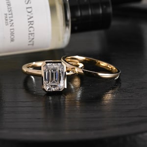 Pode incluir: Um conjunto de anéis de ouro com uma pedra central de corte esmeralda em engaste de bisel e uma banda de ouro lisa. Os anéis estão posicionados em frente a uma garrafa com um rótulo que diz Christian Dior Paris S D'Argent, acabamento polido.