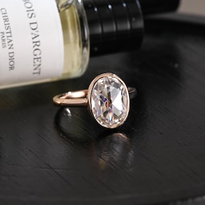 May include: A rose gold ring featuring a large, oval-cut, clear faceted gemstone set in a smooth bezel setting. The ring has a polished, slender band and is positioned near a glass bottle labelled Bois D'Argent Christian Dior Paris.