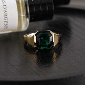 May include: A gold ring featuring a large, emerald-cut, deep green gemstone set in a polished metal band. The ring has a smooth, wide shank and a bezel setting that secures the rectangular stone, creating a bold and reflective appearance.