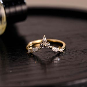 May include: A delicate gold ring with a V-shaped band adorned with small, clear gemstones. The ring features a pointed design at the top, creating a crown-like appearance.