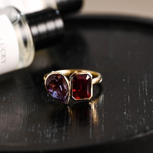 Könnte beinhalten: Ein goldener Ring mit zwei Edelsteinen. Der größere Stein ist ein quadratischer, roter Rubin. Der kleinere Stein ist ein birnenförmiger, violetter Amethyst.