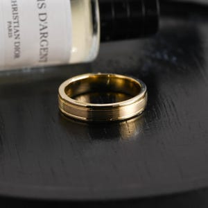 Könnte beinhalten: Ein goldfarbener Bandring mit einer gebürsteten, matten Oberfläche in der Mitte und polierten, reflektierenden Kanten. Der Ring hat ein glattes, kreisförmiges Profil mit gleichbleibender Breite und zeigt einen metallischen Glanz sowie klare Linien.
