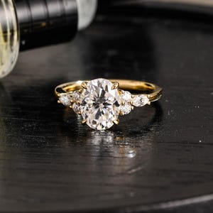 Pode incluir: Um anel de ouro com uma grande pedra preciosa transparente central de corte oval presa por garras. A banda é acentuada em cada lado por um grupo de pedras transparentes menores de corte redondo e marquise, criando um design simétrico.
