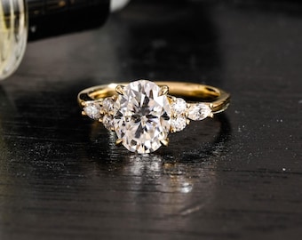 May include: A gold ring featuring a large oval-cut centre stone flanked by three smaller stones on each side, including two round-cut stones and one marquise-cut stone, set on a polished gold band that reflects light across its surface.