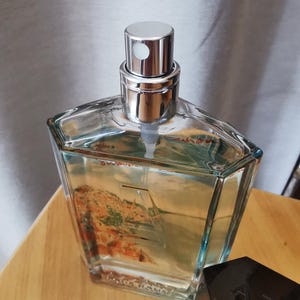 Pu&ograve; includere: Un flacone di profumo trasparente e geometrico con tappo argentato e vaporizzatore. Il flacone contiene un liquido di colore chiaro e presenta un disegno paesaggistico. Un flacone di profumo nero e angolare &egrave; accanto su una superficie di legno.