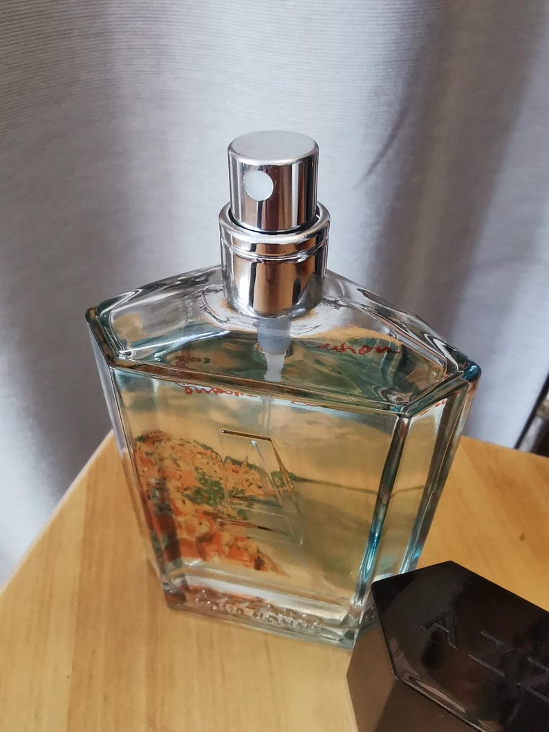 Pu&ograve; includere: Un flacone di profumo trasparente e geometrico con tappo argentato e vaporizzatore. Il flacone contiene un liquido di colore chiaro e presenta un disegno paesaggistico. Un flacone di profumo nero e angolare &egrave; accanto su una superficie di legno.