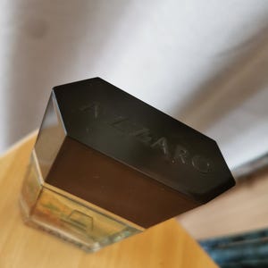 Pu&ograve; includere: Un flacone di profumo rettangolare con un tappo nero. La parola "AZZARO" &egrave; impressa sul tappo. Il flacone &egrave; parzialmente riempito con un liquido chiaro e poggia su una superficie di legno.