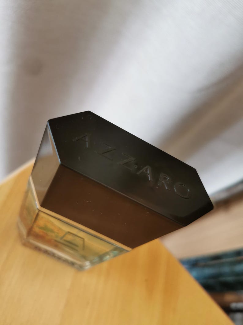 Pu&ograve; includere: Un flacone di profumo rettangolare con un tappo nero. La parola "AZZARO" &egrave; impressa sul tappo. Il flacone &egrave; parzialmente riempito con un liquido chiaro e poggia su una superficie di legno.
