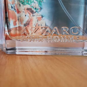 Pu&ograve; includere: Flacone di profumo in vetro trasparente con la scritta in rilievo "AZZARO POUR HOMME". Il flacone ha una forma rettangolare e un liquido azzurro all'interno. Lo sfondo mostra un'immagine sfocata di un dipinto.