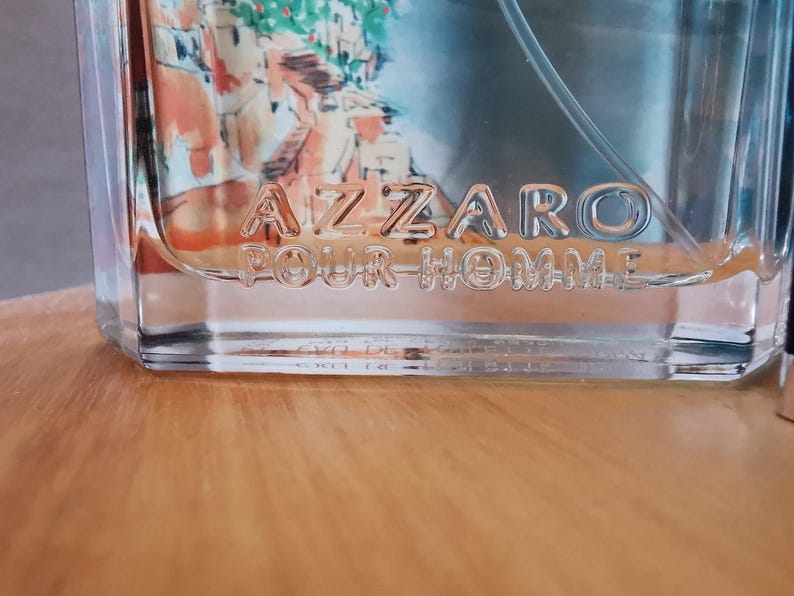 Pu&ograve; includere: Flacone di profumo in vetro trasparente con la scritta in rilievo "AZZARO POUR HOMME". Il flacone ha una forma rettangolare e un liquido azzurro all'interno. Lo sfondo mostra un'immagine sfocata di un dipinto.