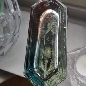 Pu&ograve; includere: Un flacone di profumo trasparente e rettangolare con una sfumatura di blu e verde. Il flacone presenta testo inciso, tra cui "EAU DE TOILETTE" e "Made in France". Il flacone &egrave; parzialmente trasparente, rivelando il liquido all'interno.