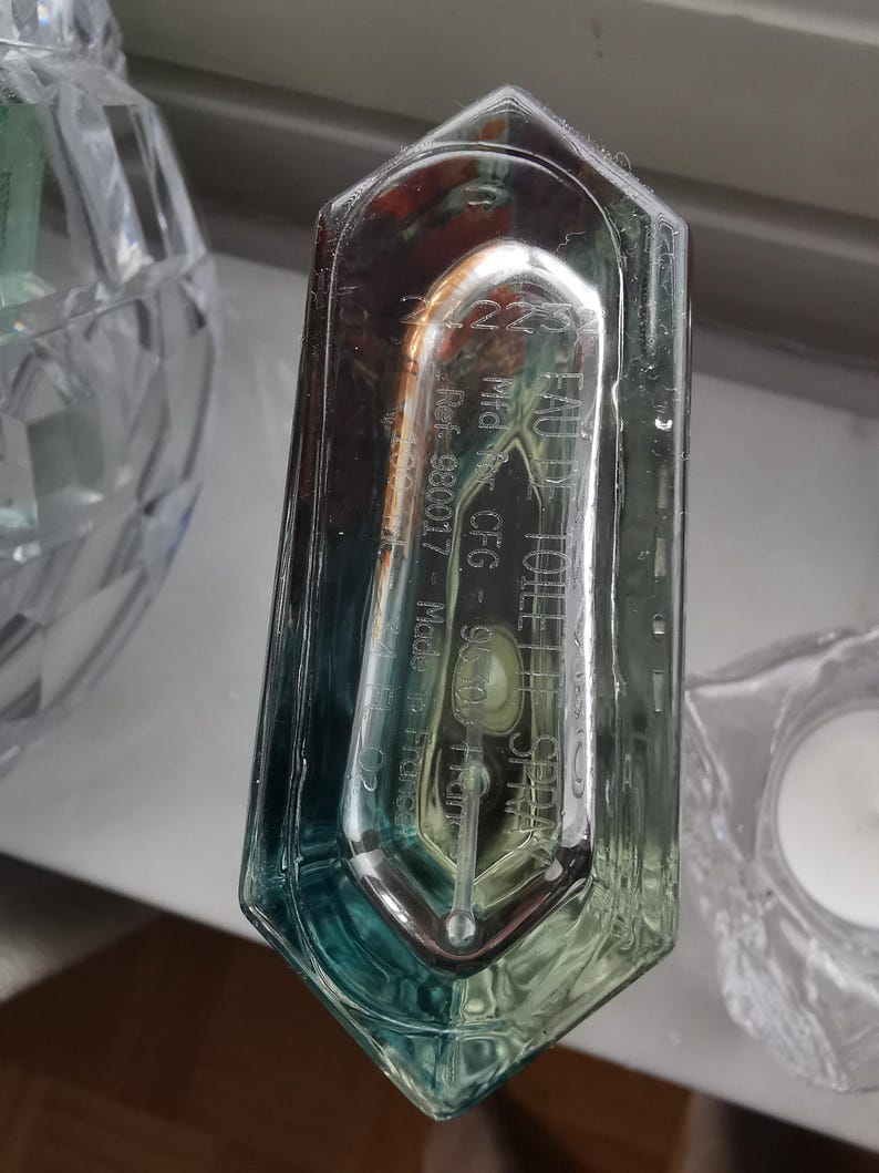 Pu&ograve; includere: Un flacone di profumo trasparente e rettangolare con una sfumatura di blu e verde. Il flacone presenta testo inciso, tra cui "EAU DE TOILETTE" e "Made in France". Il flacone &egrave; parzialmente trasparente, rivelando il liquido all'interno.