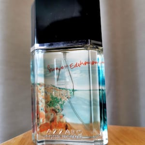 Pu&ograve; includere: Flacone di profumo in vetro trasparente con tappo nero. Il flacone presenta un'immagine panoramica di un paesaggio costiero. Il testo "Summer Edition" e "Azzaro Pour Homme" sono visibili.