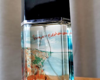 May include: A clear glass perfume bottle with a black cap. The bottle has a scenic image of a coastal landscape. The text "Summer Edition" and "Azzaro Pour Homme" are visible on the bottle.