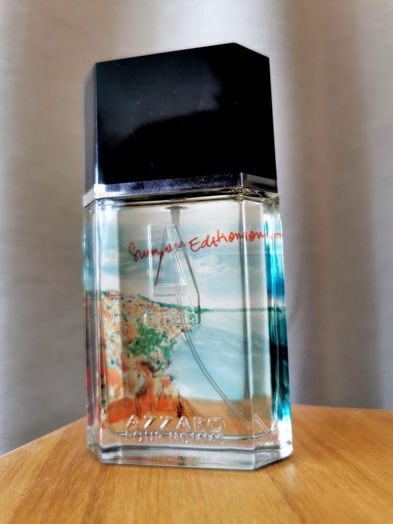 Pu&ograve; includere: Flacone di profumo in vetro trasparente con tappo nero. Il flacone presenta un'immagine panoramica di un paesaggio costiero. Il testo "Summer Edition" e "Azzaro Pour Homme" sono visibili.