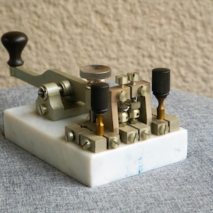 May include: A vintage telegraph key, with a black knob on a lever and two black cylindrical knobs. The key is made of metal and mounted on a white marble base. A classic piece of communication equipment.