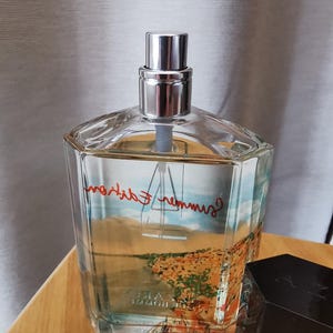 Pu&ograve; includere: Un flacone di profumo in vetro trasparente con tappo argentato. Il flacone presenta un disegno paesaggistico con le parole "Summer Edition" in rosso. Il flacone &egrave; appoggiato su una superficie di legno.
