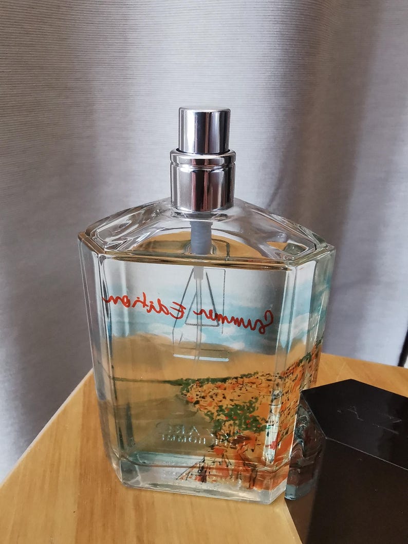 Pu&ograve; includere: Un flacone di profumo in vetro trasparente con tappo argentato. Il flacone presenta un disegno paesaggistico con le parole "Summer Edition" in rosso. Il flacone &egrave; appoggiato su una superficie di legno.