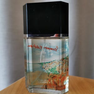 Pu&ograve; includere: Un flacone di profumo rettangolare con tappo nero. Il flacone presenta un'immagine di una citt&agrave; costiera e le parole "Summer Edition" in rosso. Il flacone &egrave; su una superficie di legno.
