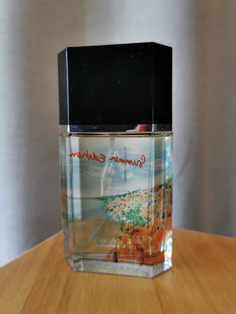Pu&ograve; includere: Un flacone di profumo rettangolare con tappo nero. Il flacone presenta un'immagine di una citt&agrave; costiera e le parole "Summer Edition" in rosso. Il flacone &egrave; su una superficie di legno.