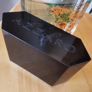 Pu&ograve; includere: Una scatola nera di forma geometrica con la scritta "AZZARO" in rilievo sulla parte superiore. La scatola &egrave; lucida e riflette la luce, appoggiata su una superficie di legno chiaro. Un contenitore di vetro &egrave; visibile sullo sfondo.