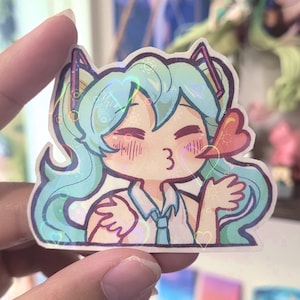 May include: A holographic sticker featuring a cartoon character with teal hair, wearing a white shirt and blue tie. The character is blowing a kiss, with the number "3" visible. The sticker has a heart-shaped bow and heart accents.