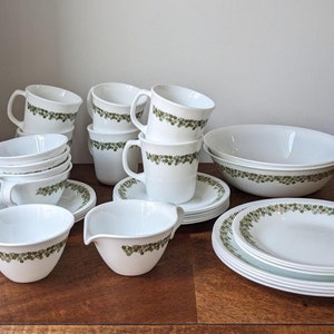 Puede incluir: Un juego de vajilla Corelle blanca con un patrón floral verde. El juego incluye tazas, platillos, una jarra de crema, un tazón y platos.