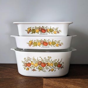 Puede incluir: Un juego de tres platos para hornear blancos con un patrón floral que incluye hojas verdes, limones amarillos, tomates rojos y especias marrones. Los platos están apilados uno encima del otro y dicen "Pechkeks" en la parte delantera.