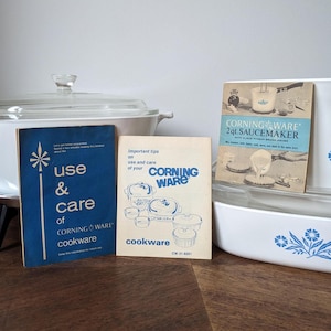 Op de afbeelding: Een verzameling Corning Ware kookgerei, waaronder een afgedekte ovenschaal, een rechthoekige bakvorm en twee instructieboekjes. De boekjes heten "Use & Care" en "2qt. SAUCEMAKER". De gerechten zijn wit met blauwe bloemmotieven.