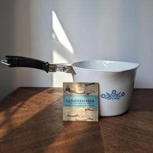 Puede incluir: Una cacerola CorningWare blanca de 1,9 litros con un asa negra y un diseño floral azul. Una tarjeta publicitaria vintage está colocada frente a la olla. La olla está sobre una superficie de madera.
