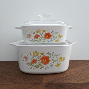 Peut inclure: Un ensemble de deux plats à gratin blancs avec des motifs floraux. Les plats présentent un motif de fleurs orange, jaunes et bleues sur fond blanc. Les plats sont empilés l'un sur l'autre.