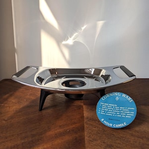 Puede incluir: Un calentador de velas Corning Ware vintage. El calentador plateado tiene un diseño curvo con asas negras y descansa sobre patas negras. Una etiqueta azul dice "Corning Ware" y "8 Hour Candle" con instrucciones.