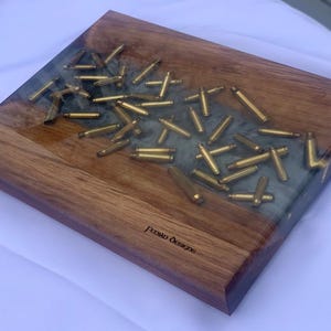 May include: A wooden cutting board with a dark wood border and a central section of clear resin embedded with numerous brass bullet casings. The board has a rectangular shape and the text "Persian Designs" is visible on the wood.