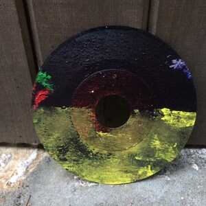 May include: A black circular disc with a hole in the center, painted with yellow, green, red, and purple paint. The disc has a textured surface.
