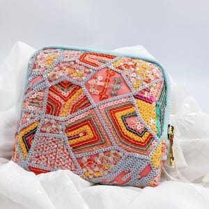 Könnte beinhalten: Eine quadratische, rosa Pailletten-Tasche mit einem geometrischen Design in Rot-, Gelb- und Orangetönen. Die Tasche ist mit einer hellblauen Paspel versehen und hat einen goldenen Reißverschlussanhänger. Das Design erinnert an Patchwork.