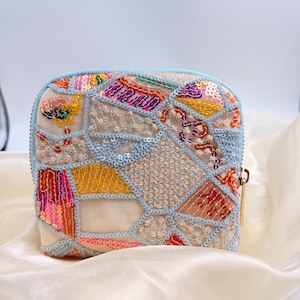 Könnte beinhalten: Eine kleine, quadratische Tasche im Patchwork-Stil mit einem hellblauen Reißverschluss und Besatz. Die Tasche ist mit bunten Pailletten und gestickten Details in Rosa, Gelb, Orange und Lila verziert. Der Hintergrund ist ein weicher, cremefarbener Stoff.